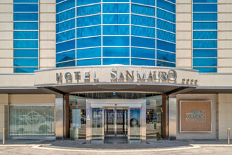 Entrance of B&B Hotel San Mauro with glass doors, beige walls, blue windows, and hotel signage above the entrance