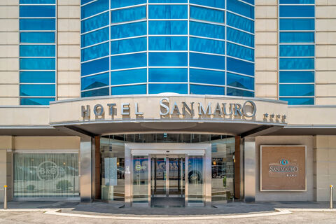 Entrance of B&B Hotel San Mauro with glass doors, beige walls, blue windows, and hotel signage above the entrance
