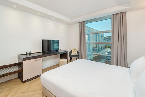 Hotel room with white bed, flat-screen TV on a desk, chair, and large window showing building view at B&B Hotel San Mauro