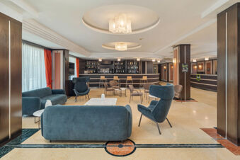 Spacious lobby area of B&B Hotel San Mauro with blue sofas, armchairs, beige chairs, bar counter, and modern lighting fixtures