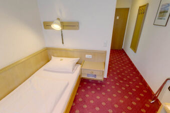 Simple hotel room with single bed, white bedding, wooden headboard, wall lamp, nightstand, and red patterned carpet at Hotel Garni Stadt Friedberg