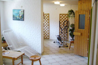 White lounge chair next to a wooden table and stool with an exercise machine and wood-paneled sauna in Hotel Garni Stadt Friedberg