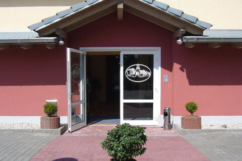 Entrance of Hotel Garni Stadt Friedberg with a red facade, double glass doors, potted plants, and a small tree in front