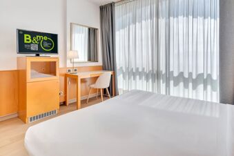 Bright hotel room at B&B Hotel Bergamo Executive featuring a bed, desk with chair and lamp, wooden cabinet, and TV with promotional screen