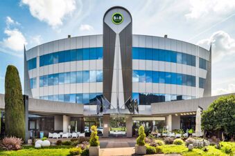 Curved gray building with blue-tinted windows, flags at entrance, greenery, and B&B Hotel Bergamo Executive logo above main door