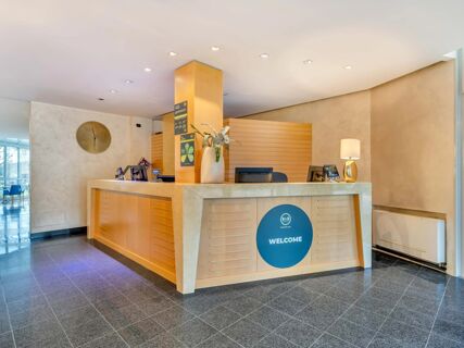 Reception desk area with wooden paneling, a large round clock, a potted plant, a lamp, and signage at B&B Hotel Bergamo Executive