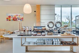 Breakfast buffet table with cereals, juice dispensers, bread, plates, cups, glasses, chafing dishes, and a toaster at B&B Hotel Bergamo Executive
