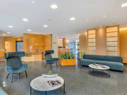 Seating area with blue armchairs, a blue sofa, marble tables, plants, and a wooden elevator area in B&B Hotel Bergamo Executive