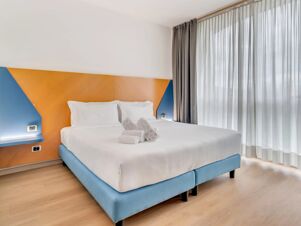A neatly made bed with white linens and towels on top, featuring a blue and orange headboard and sheer curtains in a B&B Hotel Bergamo Executive room