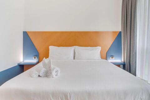 White bed with two pillows and three rolled towels on it, blue and orange headboard, grey curtains at B&B Hotel Bergamo Executive