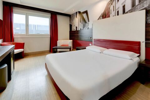 Hotel room at B&B Hotel Trento with a double bed, red and white chairs, wooden flooring, and a large window with red curtains