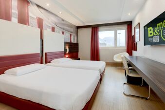Twin beds with red leather headboards, a window with red curtains, a wall-mounted TV, desk, and chairs in B&B Hotel Trento room