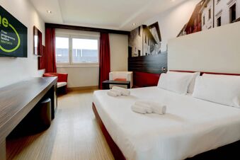 Hotel room at B&B Hotel Trento with double bed, folded towels, two armchairs, desk, large window, and wall art of buildings