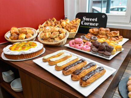 Assorted pastries including croissants, eclairs with chocolate and cream, donuts, muffins, and tart displayed on a wooden table at B&B Hotel Trento