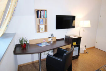 A dark wooden desk with a small plant, books on a wall shelf, flat screen TV, lamp, and black chair in Hotel G&auml;stehaus Centro Albert GmbH room