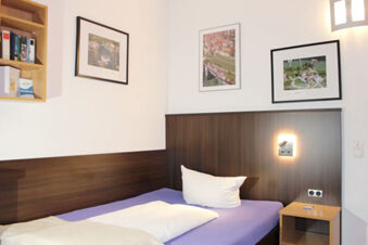 Single bed with white pillow and blanket, wooden headboard and nightstand, wall-mounted shelf and framed pictures at Hotel G&auml;stehaus Centro Albert GmbH