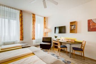 Hotel G&auml;stehaus Centro Albert GmbH room with twin beds, two windows with curtains, desk, chairs, armchair, and wall-mounted TV