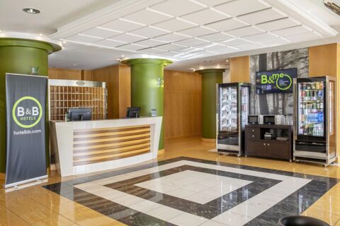 Reception area with a white and wood desk, green columns, vending machines, and B&B Hotel Cuneo Cristal signage displayed on screen and banner