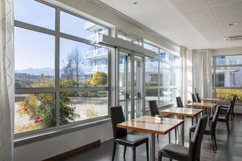 Dining area of B&B Hotel Cuneo Cristal with wooden tables, black chairs, large windows, and outdoor view of plants and buildings