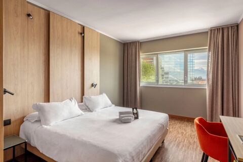 Double bed with white linens and pillows, wooden headboard, window with curtains, orange chair, and towels at B&B Hotel Cuneo Cristal