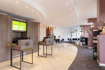 Reception area of B&B Hotel Sassuolo with two wooden kiosks, tiled floor, lounge seating, and snack shelves in a modern design