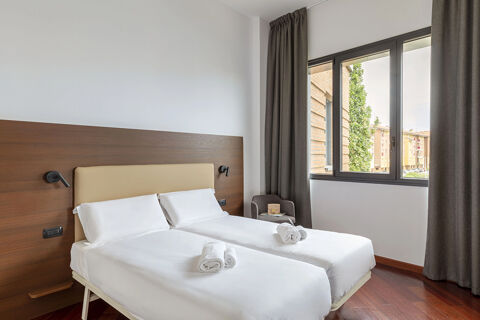 A twin bed hotel room with white linens, rolled towels, wooden flooring, dark curtains, and a window at B&B Hotel Sassuolo