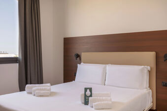 A neatly made bed with white sheets and pillows, white rolled towels on it, a wooden headboard, and a gray curtain at B&B Hotel Sassuolo