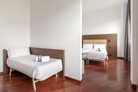 Two single beds with white linens separated by a partial wall in a room at B&B Hotel Sassuolo with wooden floors and curtains
