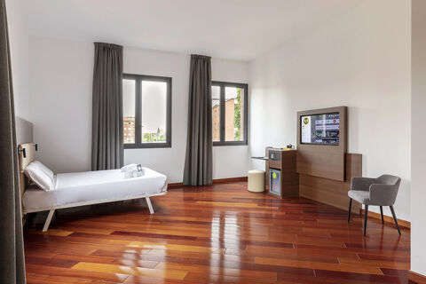 Minimalist hotel room at B&B Hotel Sassuolo with single bed, two windows with gray curtains, wooden floor, and mounted TV