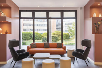Minimalist lobby seating area with orange sofa, black armchairs, small round stools, and large windows at B&B Hotel Sassuolo