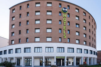 Large curved multi-story brick building with many windows and green B&B Hotel Sassuolo sign on white lower facade and entrance