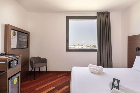 Hotel room at B&B Hotel Sassuolo featuring a white bed with towels, a wooden floor, a chair, TV, safe, mini fridge, and a window with curtains