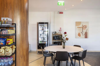 A round white table with a vase and flowers in a room with black chairs, snack shelves, and a refrigerator at B&B Hotel Sassuolo