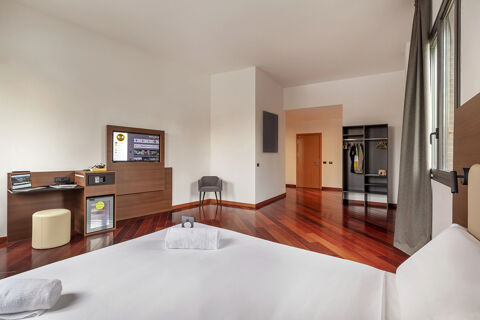 Modern hotel room at B&B Hotel Sassuolo with a large bed, wooden floor, a desk, TV, chair, and open wardrobe with clothes and hangers