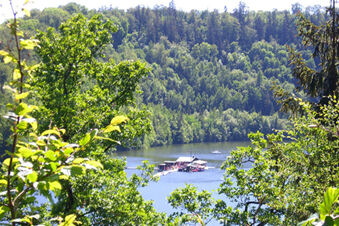 Green trees frame a lake with a floating building and a sailboat surrounded by forest at Pension K&ouml;nigshof