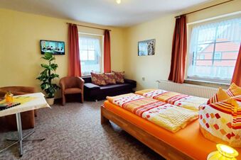 Bedroom of Pension K&ouml;nigshof with double bed, orange bedding, two windows with curtains, armchairs, small table, and sofa