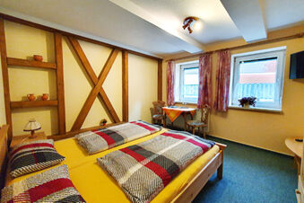 Bedroom with wooden bed and colorful bedding, small table and chairs, two windows with curtains in Pension K&ouml;nigshof