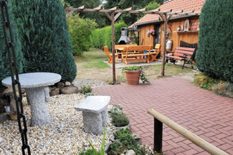 Outdoor seating area at Pension K&ouml;nigshof with stone table and bench, wooden dining set, and surrounding greenery and trees