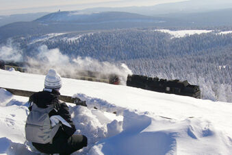 Person in winter clothing sitting on a snowy hill watching a steam train travel through a snow-covered forest near Pension K&ouml;nigshof