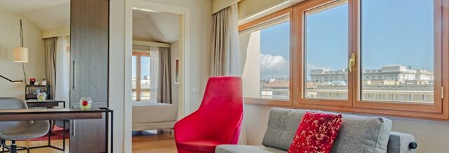 Bright hotel room at NH Collection Roma Giustiniano with a red chair, gray couch, large windows, and a view of buildings outside