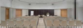 Conference room with beige chairs arranged in rows, a table with flowers, a flip chart, and wooden doors at NH Collection Roma Giustiniano