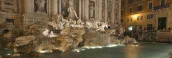 Large illuminated stone fountain with statues and cascading water at night near NH Collection Roma Giustiniano