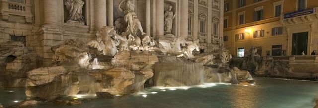 Large illuminated stone fountain with statues and cascading water at night near NH Collection Roma Giustiniano