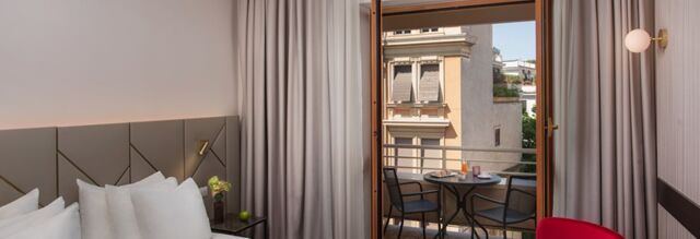 Hotel room interior at NH Collection Roma Giustiniano with a bed, pillows, bedside table, open door to balcony with table and chairs