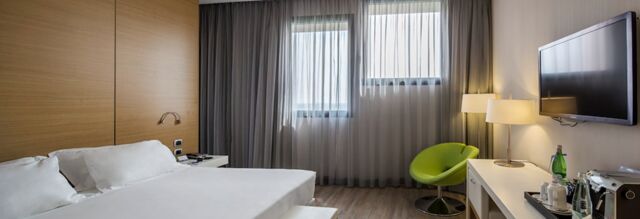 Hotel room at NH Milano Fiera with a double bed, wooden accent wall, sheer curtains, green chair, and wall-mounted TV