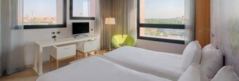 Hotel room at NH Milano Fiera with two single beds, a green chair, desk with TV, and large windows showing city views