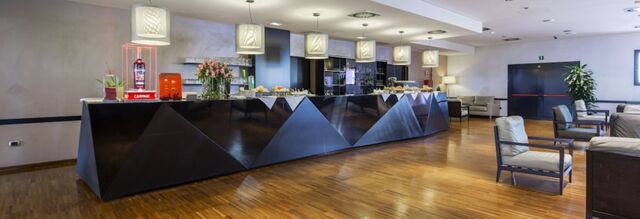 Reception area of NH Milano Fiera with a long black counter, pendant lights above, wooden floor, and seating area with armchairs and sofas
