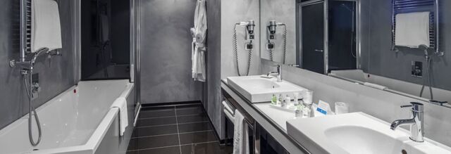 Modern bathroom at NH Milano Fiera with double sinks, large mirror, bathtub, towels, and wall-mounted hairdryers visible