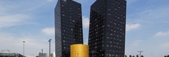Two tall black modern buildings with many windows stand behind a shorter cylindrical golden structure near NH Milano Fiera hotel