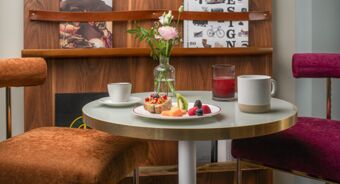 A small round table with a vase of pink flowers, a plate of fruit and toast, a white cup, glass of red juice, and a mug inside Avani Avani Palazzo Moscova Milan Hotel